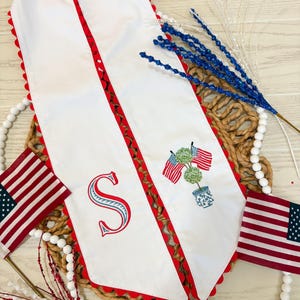 May include: White table runner with red scalloped edges and a red and brown patterned border. Features an embroidered letter "S" and a potted topiary with American flags. Includes small American flags and decorative accents.