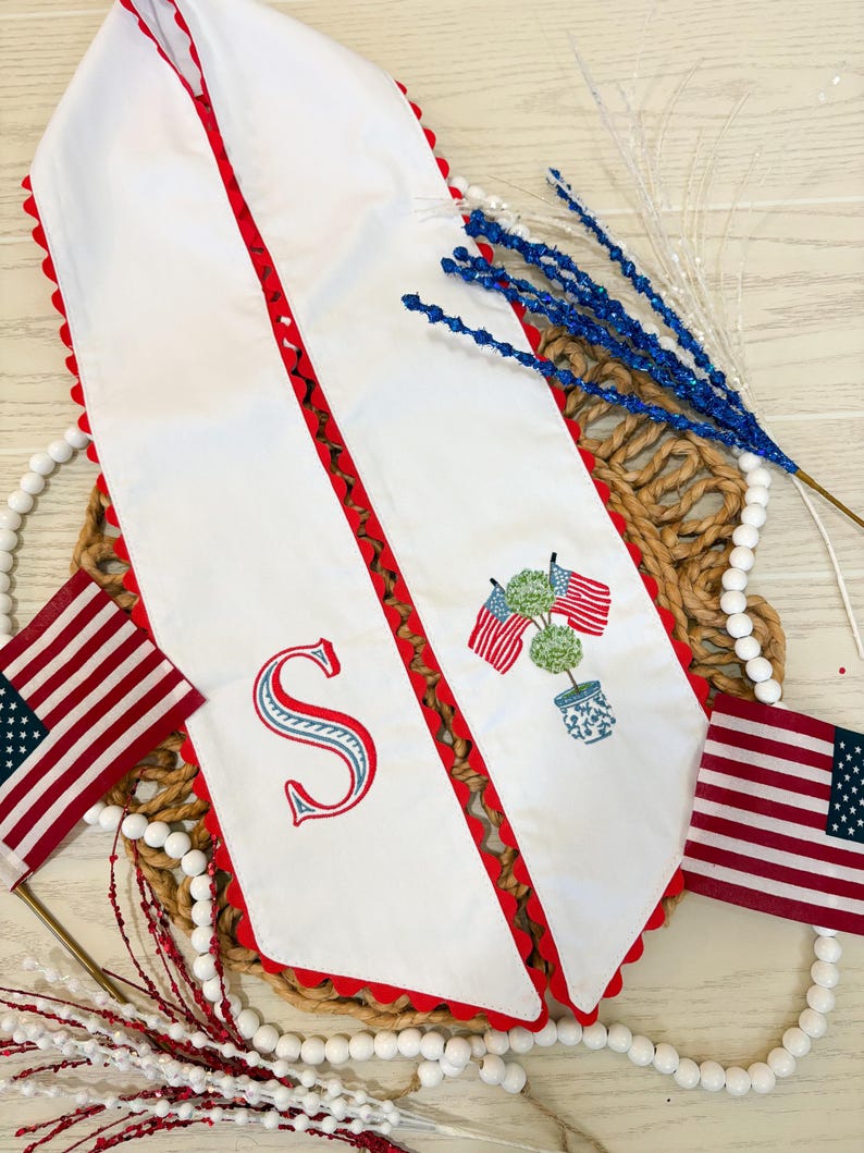 May include: White table runner with red scalloped edges and a red and brown patterned border. Features an embroidered letter "S" and a potted topiary with American flags. Includes small American flags and decorative accents.