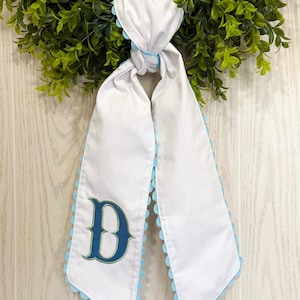 Könnte beinhalten: Weißes Stoffband mit hellblauem, gewelltem Rand und einem großen, mit Teal bestickten Buchstaben "D". Das Band ist verknotet und hängt vor einem grünen Kranz. Der Hintergrund ist eine helle Holzmaserung.