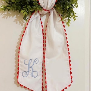 May include: White fabric sash with red scalloped edges and a light blue embroidered letter "K". The sash is tied to a green wreath, creating a decorative accent. The design is simple and elegant.