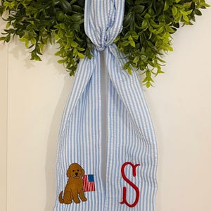 Puede incluir: Un colgador de puerta a rayas azules y blancas con un perro marrón sosteniendo una bandera estadounidense y una letra "S" roja bordada. El colgador de puerta cuelga de una corona verde.