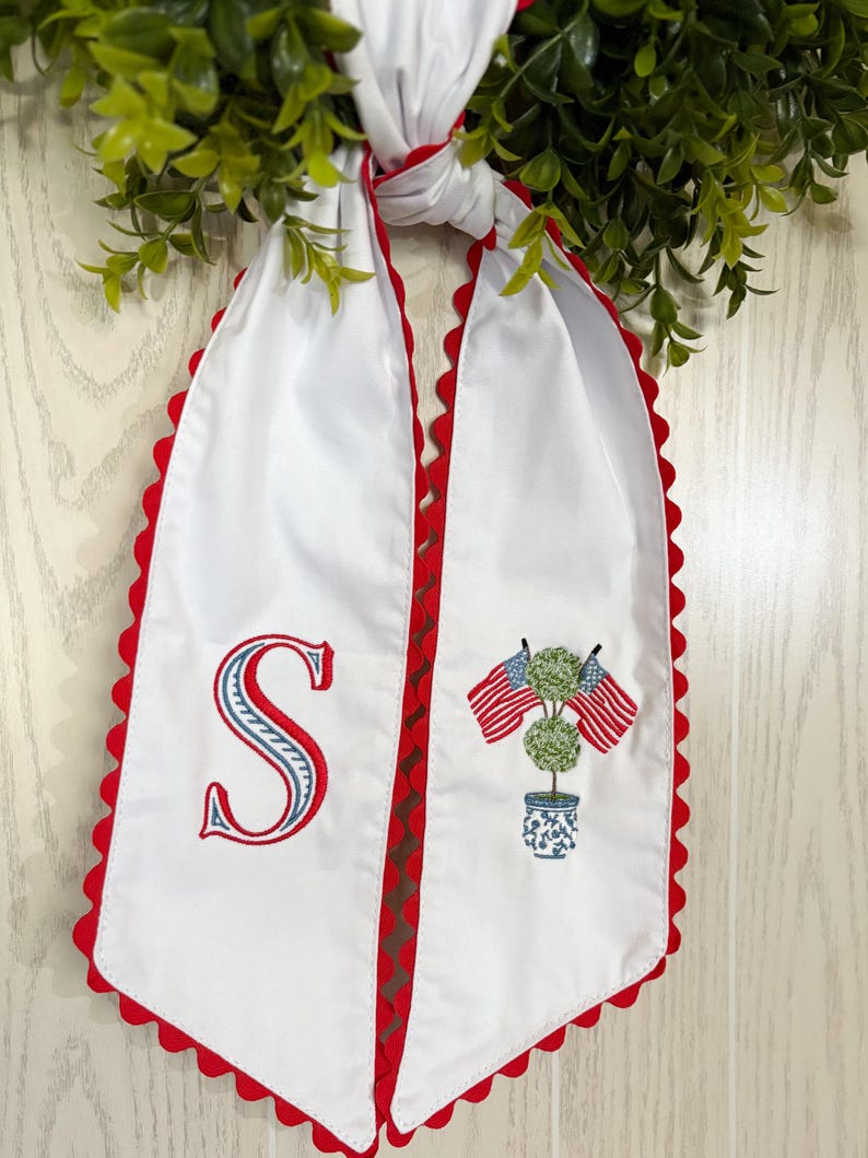 May include: White fabric sash with red scalloped edges. Features an embroidered letter "S" and a design of two American flags and a topiary in a blue and white pot. The sash is tied to a green wreath.