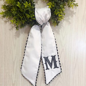 May include: A white satin wreath sash with a black scalloped edge and a large, embroidered gray letter "M". The sash is tied to a green wreath with a white satin knot.