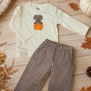 May include: A long-sleeve cream-colored shirt featuring an embroidered brown and white checkered puppy sitting inside an orange polka-dot pumpkin, paired with brown and white checkered pants with an elastic waistband for a complete outfit.