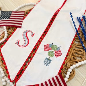 May include: White fabric table runner with red scalloped edges and embroidered patriotic designs. Features an embroidered letter "S", American flags, and a topiary in a blue and white pot. Decorative accents include small American flags.