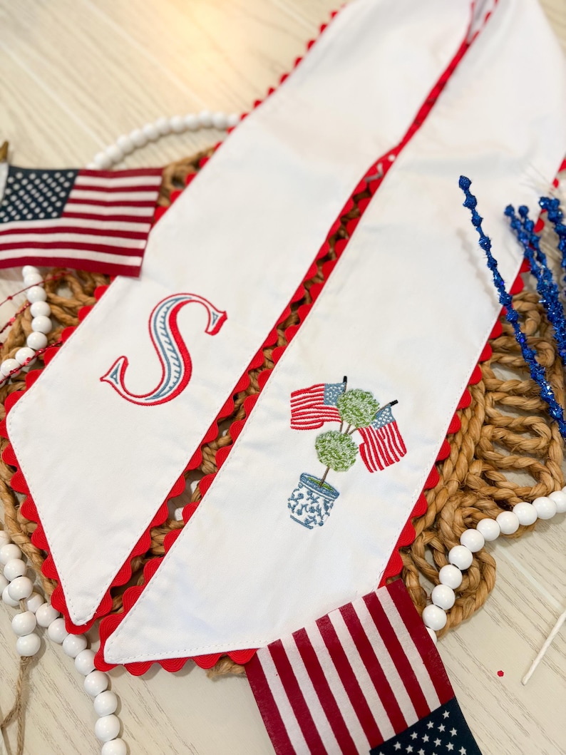 May include: White fabric table runner with red scalloped edges and embroidered patriotic designs. Features an embroidered letter "S", American flags, and a topiary in a blue and white pot. Decorative accents include small American flags.
