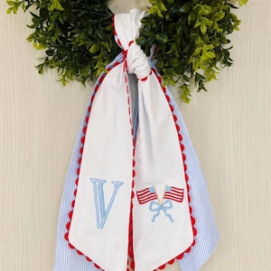 May include: A decorative wreath with a green leafy base, adorned with a white and blue fabric sash. The sash features a large embroidered blue letter "V" and a design of two American flags. The edges are trimmed with red scalloped detailing.