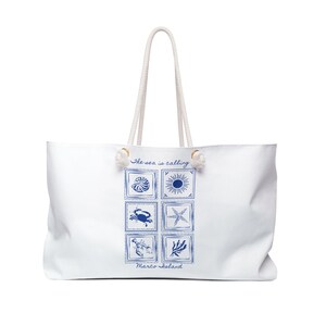 Op de afbeelding: Witte draagtas met een touwhandvat. De tas heeft een blauw vierkant ontwerp met illustraties met een zeethema, waaronder een zeeschelp, een zon, een krab, een zeester en zeewier. De tekst "The sea is calling" en "Marco Island" staan ook op de tas.