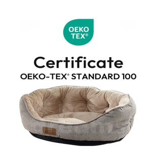 Op de afbeelding: Een hondenbed met een grijze buitenkant en een zacht, beige interieur. Het bed heeft een ronde vorm met een zwarte basis. De afbeelding bevat de tekst "Certificate OEKO-TEX&reg; STANDARD 100" en het OEKO-TEX-logo.