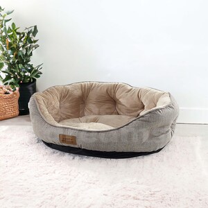 Op de afbeelding: Ovaalvormig hondenbed met een grijze stoffen buitenkant en een zacht, beige interieur. Het bed heeft een zwarte basis en een klein, bruin label. Het staat op een wit, pluizig tapijt, met een plant en een mand op de achtergrond.