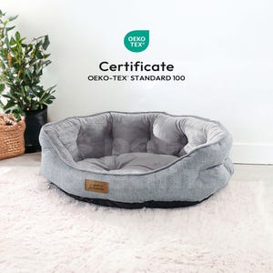 Op de afbeelding: Een grijs huisdierbed met een zacht interieur en een textuur aan de buitenkant. Het bed heeft een ronde vorm en een donkere basis. De tekst "Certificate OEKO-TEX&reg; STANDARD 100" is inbegrepen.