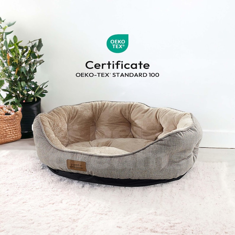 Op de afbeelding: Een grijs en beige huisdierbed met een zacht interieur en een zwarte basis. Het bed heeft een ovale vorm en een klein label. De tekst "OEKO-TEX&reg; Certificate STANDARD 100" is ook aanwezig.