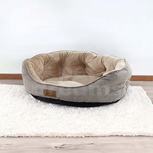 Op de afbeelding: Ovaal huisdierbed met een grijze stoffen buitenkant en een zachte, bruinachtige binnenkant. Het bed staat op een wit, pluizig kleed. Een klein, bruin label met tekst staat aan de zijkant van het bed.