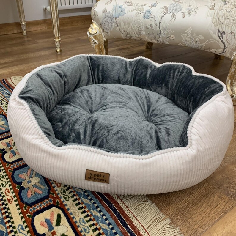 May include: A round pet bed with a white corduroy exterior and a plush, gray interior. The bed features a soft, cushioned center and a raised edge for comfort. A small, rectangular tag with the text "pet dreams" is sewn on the side.