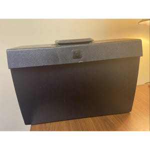 May include: A black rectangular storage box with a textured lid and a handle. The box has a clasp on the front and is sitting on a wooden surface.