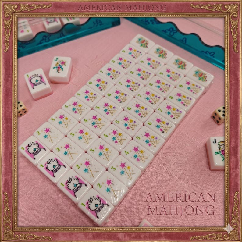 May include: A set of white American Mahjong tiles featuring colorful illustrations of flowers, stars, and numbered characters. The tiles are arranged in a grid pattern with additional loose tiles and two dice visible. The text AMERICAN MAHJONG is printed on the image.