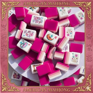 May include: A collection of American Mahjong tiles featuring a vibrant pink and white color scheme. The tiles display various colorful illustrations including candy, stars, a crescent moon, and a face, alongside standard numerical and letter markings.