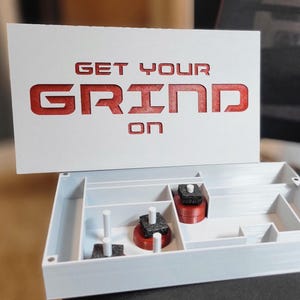May include: A white box with the words "GET YOUR GRIND ON" in red. The box is open, revealing compartments with red and black objects inside. The design is modern and minimalist.