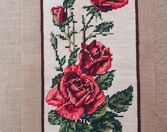 May include: Needlepoint tapestry featuring a vertical arrangement of red roses with green leaves and stems. The roses are in various stages of bloom, set against a cream-colored background with a brown border. The needlepoint is on a beige canvas.