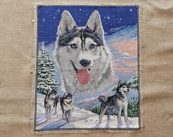 May include: Cross-stitch embroidery of a husky dog with blue eyes and a pink tongue. The husky is surrounded by a snowy landscape with a forest in the background. Three smaller husky dogs are also depicted in the scene.
