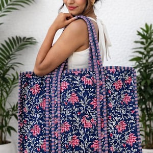 May include: A large rectangular quilted tote bag featuring a dark blue background with a repeating pattern of pink flowers and white vines. The bag has two long shoulder straps and edges trimmed with a blue and white striped fabric pattern.