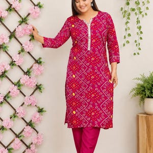 May include: A magenta knee-length tunic featuring a white and yellow geometric tie-dye pattern, three-quarter sleeves, and a silver sequined trim along the neckline, paired with matching solid magenta trousers.