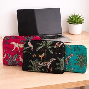 May include: Three laptop sleeves with vibrant jungle animal patterns. The sleeves are red, black, and teal, each displaying a unique design of zebras, cheetahs, and birds amidst tropical foliage. A laptop and a potted plant are in the background.
