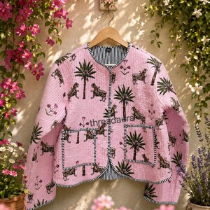 Puede incluir: Chaqueta acolchada rosa con estampado de tigre y palmeras. La chaqueta tiene forro y ribete a rayas blancas y negras. Cuelga de una percha de madera sobre un fondo floral.