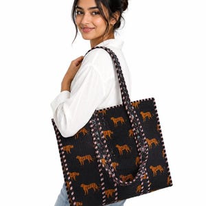 May include: A large, rectangular, quilted tote bag in black, featuring a repeating pattern of orange and black cheetahs. The bag has two shoulder straps with a striped black and white pattern, and matching striped trim along the edges.