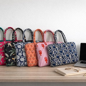 Op de afbeelding: Een verzameling kleurrijke tote bags met diverse patronen, waaronder bloemen, tropische en gestreepte ontwerpen. De tassen staan op een houten oppervlak naast een laptop, notitieboek en bril. Er staat ook een kleine plant in een pot.