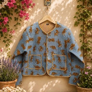 May include: A light blue quilted jacket with a tiger print. The jacket has two front pockets and a yellow and white striped lining. The trim is a gold color. The jacket is hanging on a wooden hanger.