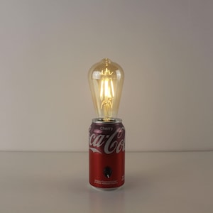 Może przedstawiać: Unikalna lampa wykonana z puszki Cherry Coca-Cola. Czerwona puszka zawiera logo i tekst Coca-Cola. Żarówka w stylu vintage znajduje się na górze, emitując ciepłe światło. Lampa posiada czarny włącznik/wyłącznik.