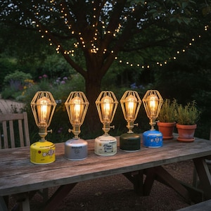 Op de afbeelding: Vijf decoratieve lampen met een vintage industrieel ontwerp, met een kooilamp en een voet die lijkt op een gaspatroon. De lampen staan op een houten tafel buiten, met lichtslingers op de achtergrond. De voeten zijn geel, zilver, wit, groen en blauw.
