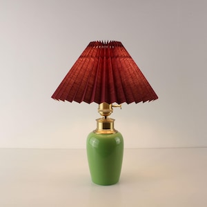 May include: A green ceramic table lamp with a pleated burgundy lampshade. The lamp has brass accents and a rounded base. The lampshade is cone-shaped and casts a warm glow. The lamp is a decorative lighting fixture.