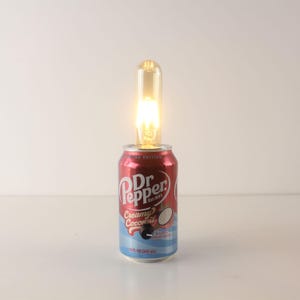May include: A unique lamp made from a Dr. Pepper Creamy Coconut soda can. The red and blue can features the Dr. Pepper logo and a coconut illustration. A glowing light bulb is inserted into the top of the can, illuminating the space.