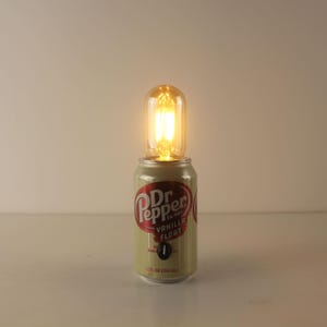Op de afbeelding: Een hergebruikte Dr. Pepper Vanilla Float-blik met een gloeiende Edison-stijl gloeilamp erop. Het blik is lichtgroen met het Dr. Pepper-logo in rood en wit. De gloeilamp is helder en geeft een warm, geel licht af.