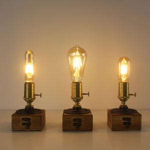 Può includere: Tre lampade da tavolo in stile vintage con lampadine Edison. Ogni lampada presenta una base in legno, un portalampada color ottone e una leva decorativa. Le lampadine sono accese, emettendo un bagliore caldo. Le lampade sono per interni.