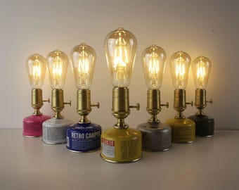 May include: A collection of vintage-style table lamps. Each lamp features a brass fixture with a glowing Edison-style bulb. The bases are repurposed camping gas canisters in various colors, including pink, blue, yellow, and black. The lamps are arranged in a row.