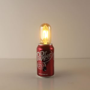 Peut inclure: Lampe originale composée d'une canette rouge Dr. Pepper surmontée d'une ampoule de style vintage. La canette porte le logo Dr. Pepper, le texte "Est. 1885" et "12 FL OZ (355 ml.)". L'ampoule diffuse une lumière chaude et accueillante.