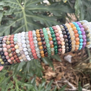 May include: A collection of beaded bracelets in various colors, including black, brown, green, pink, and blue. The bracelets are made of small, round beads and are displayed on a white cylindrical object. The background features green foliage.