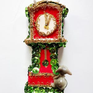 Puede incluir: Adorno navideño decorativo con forma de reloj de pie. El reloj tiene una esfera roja con adornos dorados y una esfera blanca. Está adornado con lentejuelas verdes y una pequeña figura. Una decoración festiva para las fiestas.