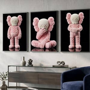 May include: Three framed art prints featuring pink and white cartoon-like figures against a black background. The figures have crossed-out eyes and are in different poses. The artwork is displayed in a modern living room setting.