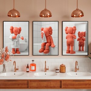 May include: Three framed wall art prints featuring a coral-colored cartoon character in a bathroom setting. The prints are displayed above a bathroom vanity with three sinks, copper fixtures, and a wooden cabinet base with white tiled walls.