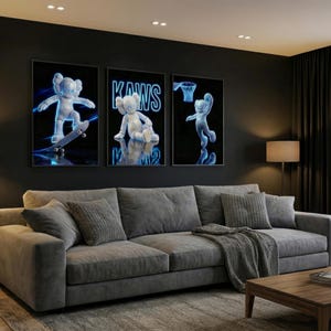 May include: Three framed art prints featuring a white cartoon figure with blue neon outlines. The prints depict the figure skateboarding, sitting, and playing basketball against a black background. The prints are above a gray sofa.
