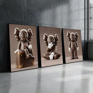 May include: Three framed art prints showcasing brown figures with X-marked eyes and hands. One figure sits on a brown suitcase, another covers its face, and the third stands. The figures have a repeating pattern.