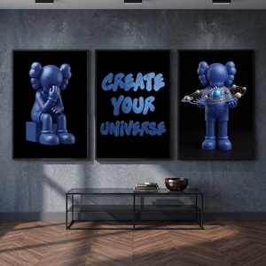 May include: A set of three framed wall art prints with a black background. The left print shows a blue cartoon character sitting. The center print features the blue text "CREATE YOUR UNIVERSE". The right print shows a blue character holding a solar system.