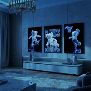 May include: Three framed artworks featuring a neon-style KAWS character in various poses: skateboarding, seated, and playing basketball. The artworks are displayed in a modern living room with blue lighting, a low cabinet, and a chandelier.