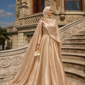 Könnte beinhalten: Ein langes, fließendes champagnerfarbenes Kleid mit passendem Umhang. Das Kleid hat lange Ärmel und einen hohen Ausschnitt, mit einer floralen Verzierung an der Schulter. Die Frau hält einen kleinen Strauß weißer Blumen. Der Hintergrund ist ein großes Gebäude mit Treppen.