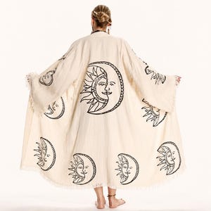 May include: Cream-colored kimono-style robe with a black sun and moon design. The robe features a fringed hem and sleeves, with the celestial pattern repeated across the garment. The robe is made of a lightweight fabric.