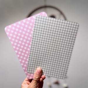 Könnte beinhalten: Zwei rechteckige Karten mit abgerundeten Ecken, eine rosa und eine graue, beide mit Vichy-Muster. Die Karten werden vor einem verschwommenen Hintergrund gehalten, der einen Traumfänger zeigt.